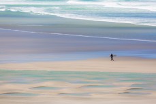 A surfer walks along a remote beach on Otago Peninsula, Dunedin, New Zealand. 